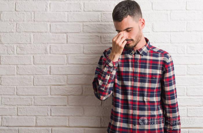 年轻的成年男子站在白色砖墙上疲惫的揉鼻子和眼睛感到疲倦和头疼.