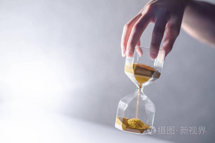 通过男性手上的现代沙漏形状的褐色沙子的关闭.时间流逝, 时间过快.