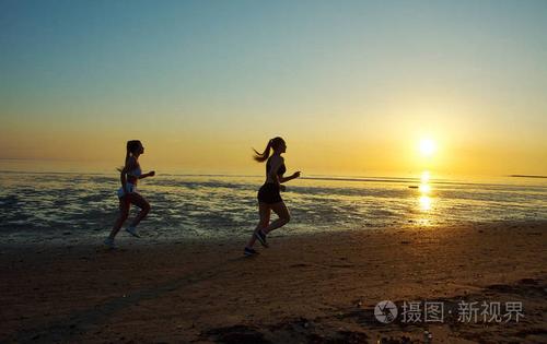 日出时两个女孩在海边奔跑的剪影,日落的黑色海滩背景