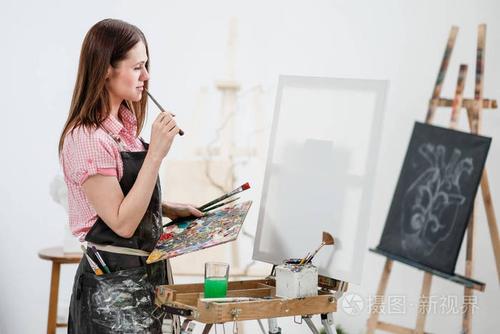 一位年轻的女画家在明亮的白色工作室里在画架上的画布上画一幅画