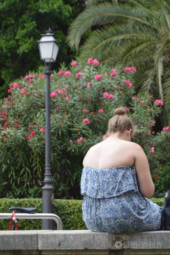 胖女孩穿着衣服,赤裸的背,疲惫的放松和日光浴,坐在公园的长凳上,在