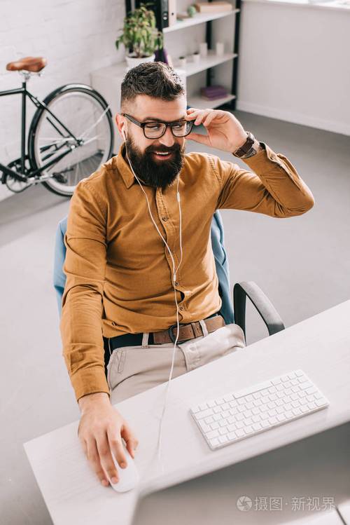 戴着耳机坐在办公室的电脑桌上工作的大胡子成年商人