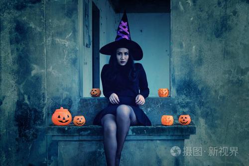 照片 穿着女巫万圣节服装的年轻女人在可怕的黑暗背景上抱着一个橙色
