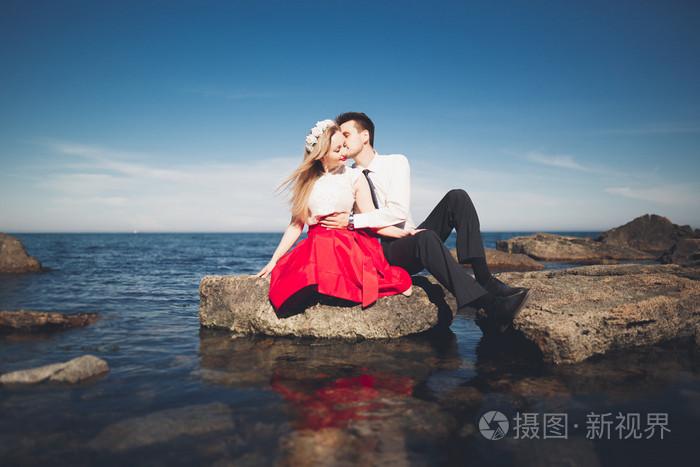 浪漫恩爱的夫妻,靠近海,蓝色天空的石头上摆姿势