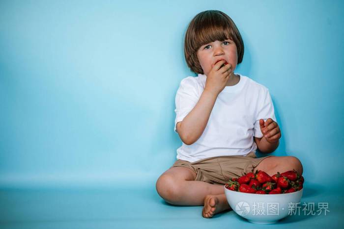 蹒跚学步的男孩坐在蓝色背景上碗里放着草莓